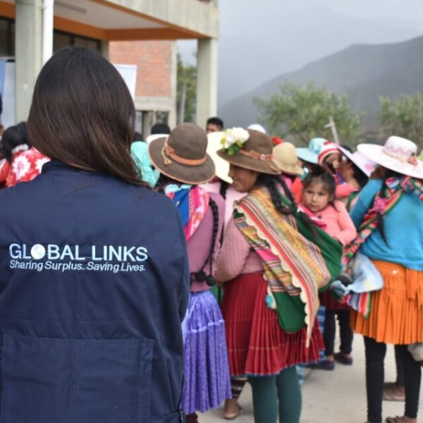 Una mujer de pie con un chaleco que dice Global Links. En el fondo hay mujeres vestidas con ropa tradicional boliviana de pie en un grupo con niños.