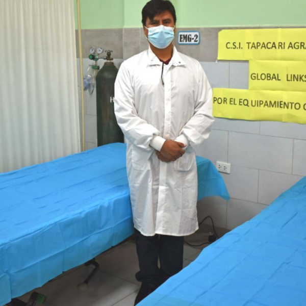 A doctor standing next to hospital beds with a white coat and mask on.